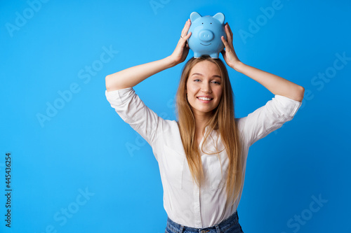 Beautiful smiling young woman with piggy bank on blue background