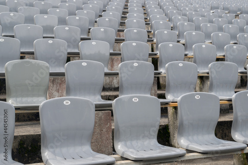 Rows of gray plastic chairs with numbers on the backs, installed in rows on cement tables in the street area, a selective focus. The front view of the seats, going into a blurry perspective.