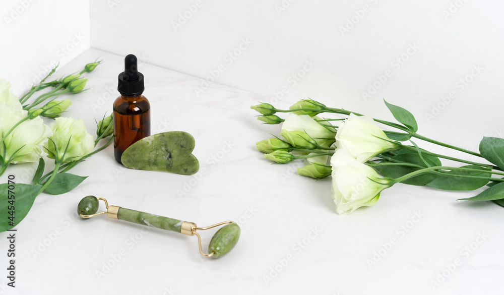 Jade roller for face massage. Green gua sha on the marble white background with flat lay. Anti age, lifting and toning treatment, acupressure. Eustoma flowers. Serum brown bottle with pipette Jade roller for face massage. Green gua sha on the marble white background with flat lay. Anti age, lifting and toning treatment, acupressure. Eustoma flowers. Serum brown bottle with pipette