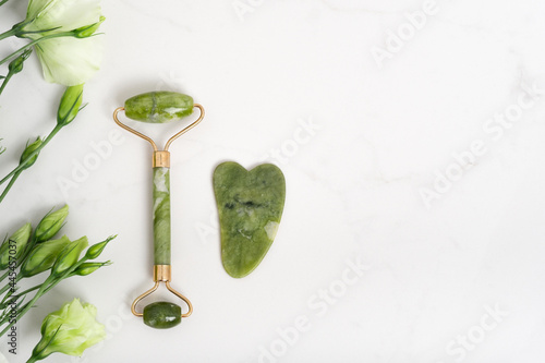 Jade roller for face massage. Green gua sha facial massager tools on the marble white background with copy space flat lay. Anti age, lifting and toning treatment, acupressure. Eustoma flowers