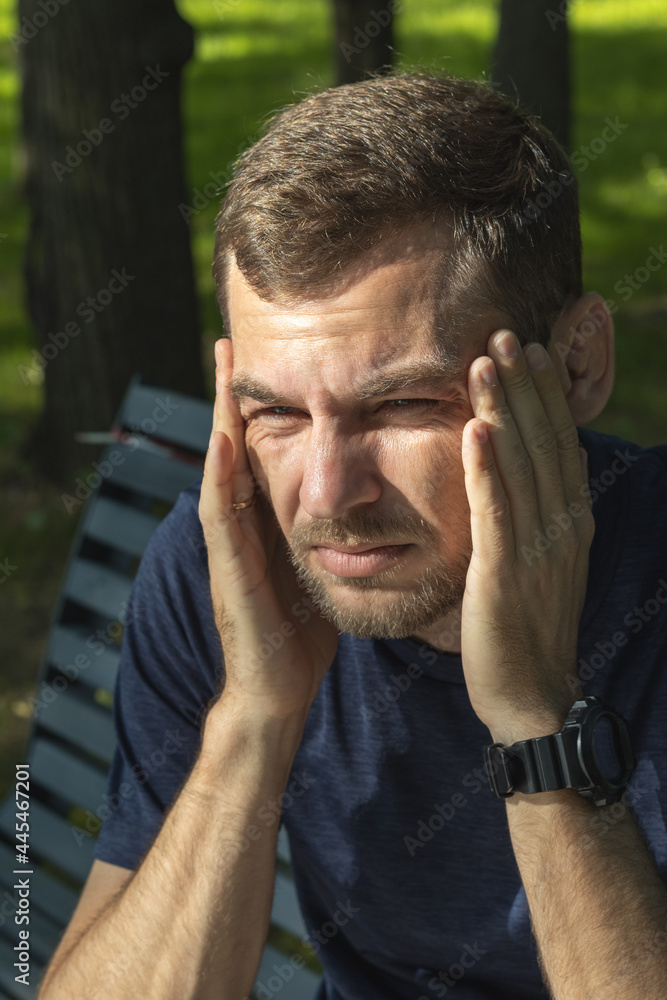 Adult man holds his head with hand while sitting on bench in park. Concept of worries, stressed, headache.