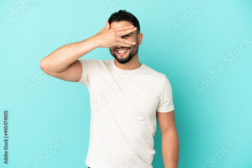 Young arab man isolated on blue background covering eyes by hands and smiling © luismolinero