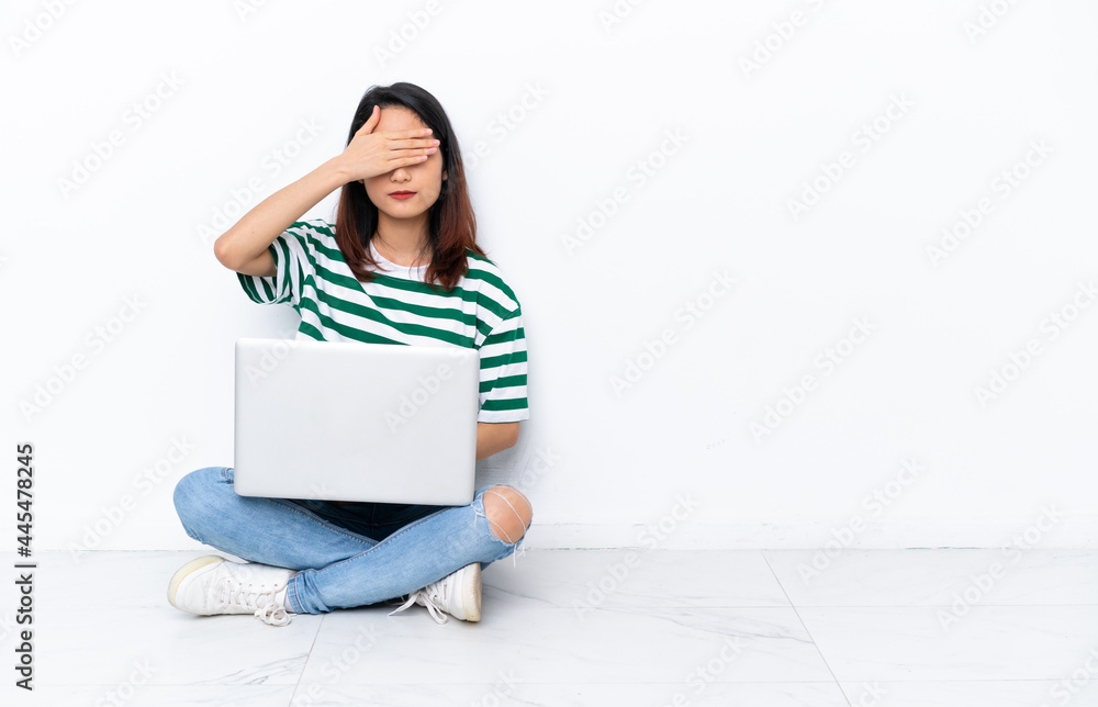 Young Vietnamese woman with a laptop sitting on the floor isolated on white wall covering eyes by hands. Do not want to see something Young Vietnamese woman with a laptop sitting on the floor isolated on white wall covering eyes by hands. Do not want to see something