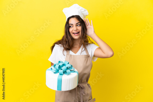 Young caucasian pastry chef woman with a big cake isolated on yellow background listening to something by putting hand on the ear