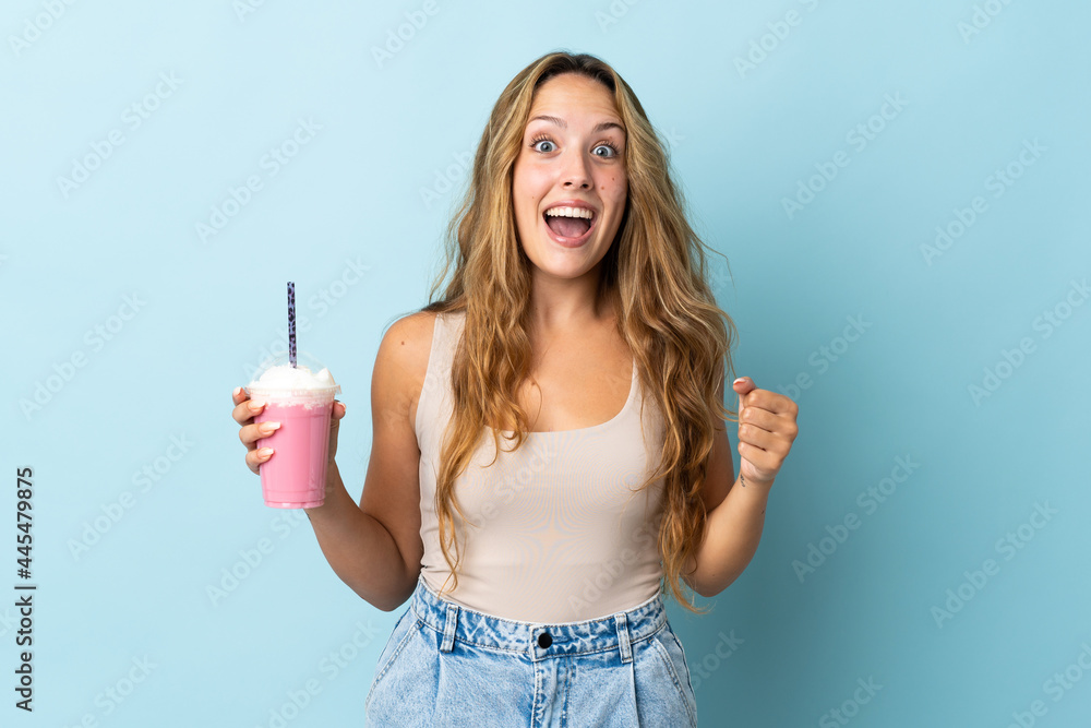 Young woman with strawberry milkshake isolated on blue background celebrating a victory in winner position Young woman with strawberry milkshake isolated on blue background celebrating a victory in winner position