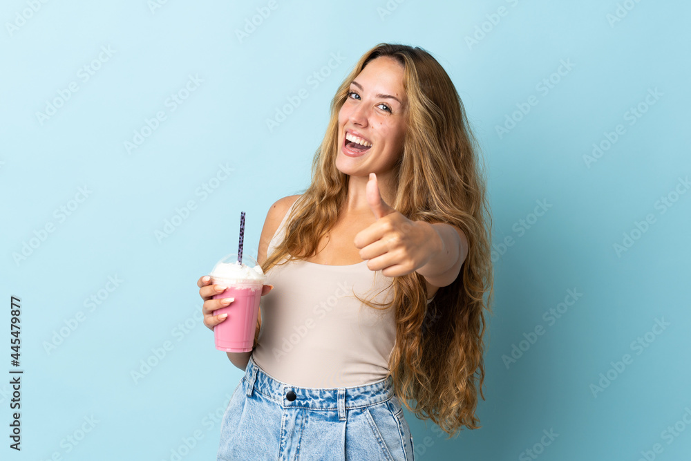 Young woman with strawberry milkshake isolated on blue background with thumbs up because something good has happened Young woman with strawberry milkshake isolated on blue background with thumbs up because something good has happened