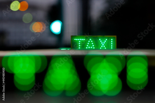 Taxi cab sign illuminated on top of vehicle at nighttime. City traffic lights with neon color in the background.