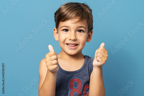 Front view of small caucasian boy four years old standing in front of blue background studio shot smiling and holding thumbs up child support and success concept