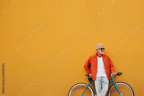 Smiling man in orange jacket and white shirt poses with bicycle. Attractive adult with white beard in light jeans posing on background of orange wall..