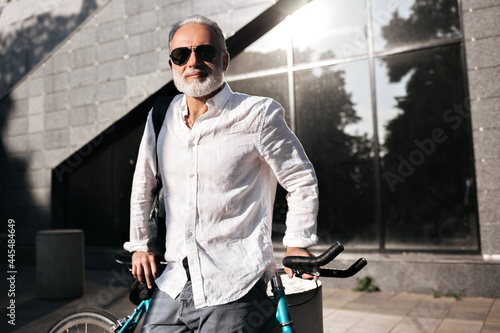 Charming man in white shirt and sunglasses poses outside with bicycle. Attractive adult guy in glasses and office clothes smiling..