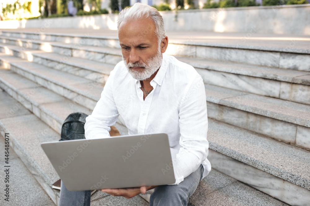 Adult man in white shirt poses with laptop. Handsome bearded guy in bright office clothes is sitting on stairs with serious face.. Adult man in white shirt poses with laptop. Handsome bearded guy in bright office clothes is sitting on stairs with serious face..