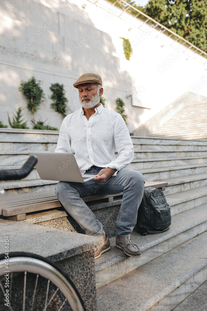 Handsome man in cap and shirt works with laptop. Modern business guy in stylish clothes with backpack sits on bench and poses.. Handsome man in cap and shirt works with laptop. Modern business guy in stylish clothes with backpack sits on bench and poses..