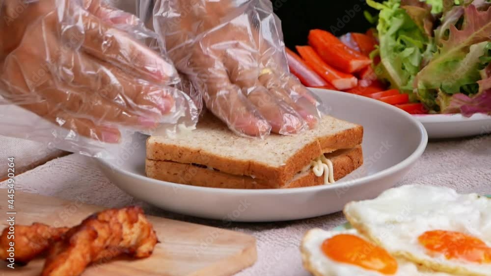 Woman in gloves cooking Whole Wheat Bread Sandwich with fried chicken, lettuce, tomato topped with mayonnaise and fried egg topped with ketchup on a plate in the kitchen. Healthy food concept. Woman in gloves cooking Whole Wheat Bread Sandwich with fried chicken, lettuce, tomato topped with mayonnaise and fried egg topped with ketchup on a plate in the kitchen. Healthy food concept.