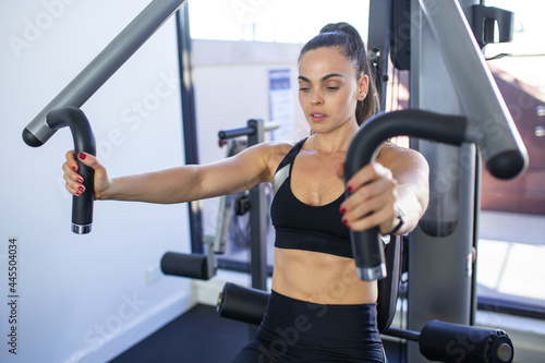 fitness woman wearing black sportswear sitting doing exercise in a machine looking down, inside of a small gym