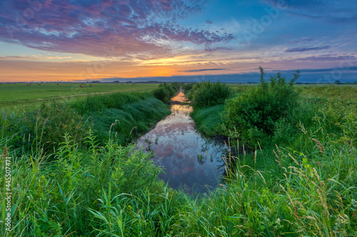 Colorful sunrise with clouds over green fields and overgrown canal, where the sky is reflected as in a mirror