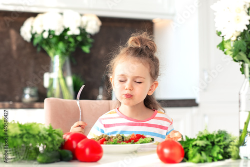 child girl enjoy eating healthy salad with closed eyes from a large plate at home in white sunny kitchen. Fresh vegetables and flowers on table.Healthy Nutrition and Vegan concept for kid