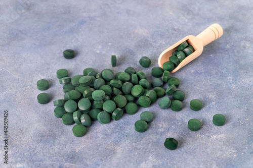 Green pills of spirulina or chlorella on a gray-blue plaster background in a glass bowl and in a wooden spoon. Healthy superfood diet and detox nutrition concept. photo