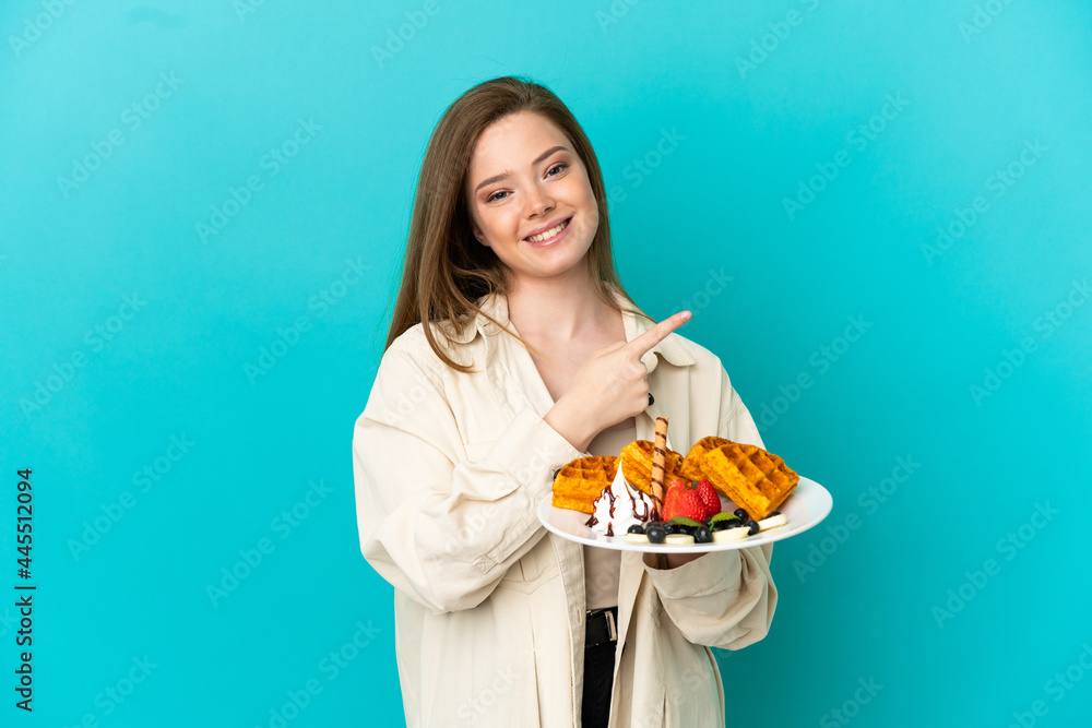 Teenager girl holding waffles over isolated blue background pointing to the side to present a product Teenager girl holding waffles over isolated blue background pointing to the side to present a product