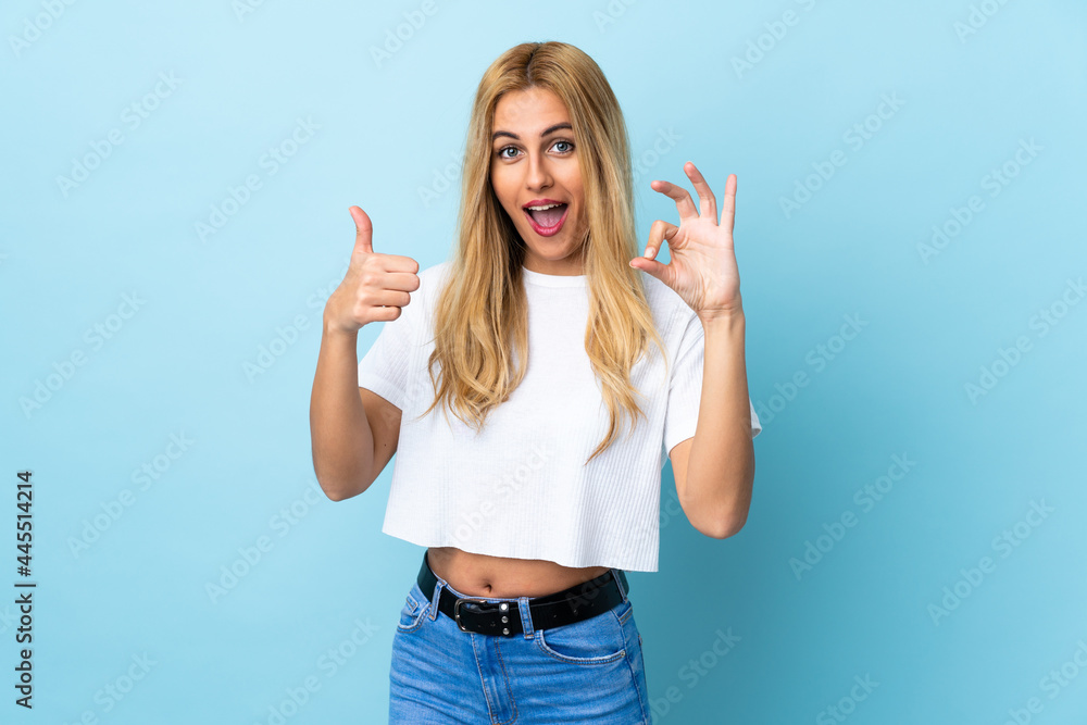 Young Uruguayan blonde woman over isolated blue background showing ok sign and thumb up gesture Young Uruguayan blonde woman over isolated blue background showing ok sign and thumb up gesture