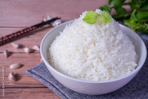 Steamed organic white rice in a white ceramic bowl with chopsticks and apron and green coriander stained on a brown wooden table.
