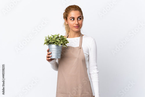 Young blonde gardener woman girl holding a plant over isolated white background standing and looking to the side