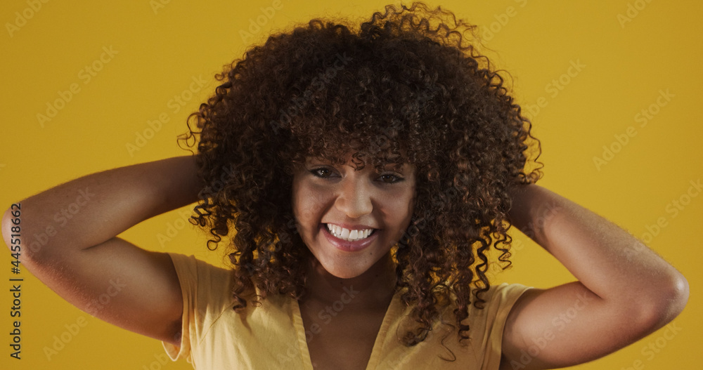 Happy laughing American African woman with her curly hair on yellow background. Laughing curly woman in sweater touching her hair and looking at the camera. Happy laughing American African woman with her curly hair on yellow background. Laughing curly woman in sweater touching her hair and looking at the camera.
