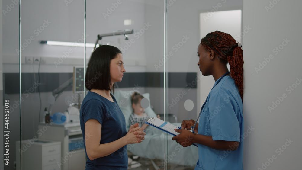 Fototapeta premium African american practitioner nurse monitoring sickness symptom explaining medical treatment to worried mother during clinical consultation. Hospitalized child resting in bed playing games on phone