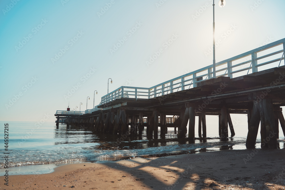 Beautiful wooden pier on the seashore. Strong sunlight during summer. Promenade place with wooden floor and painted in white handrails over the Baltic Sea.