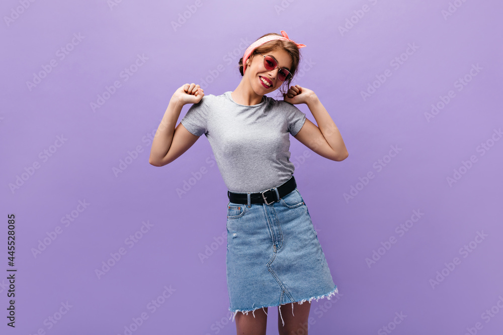 Lady in good mood poses on purple background. Charming young woman in pink summer sunglasses, gray t-shirt and denim skirt smiling.. Lady in good mood poses on purple background. Charming young woman in pink summer sunglasses, gray t-shirt and denim skirt smiling..