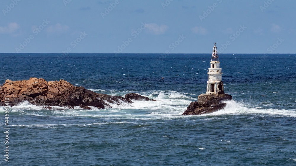 Attractive old lighthouse seascape, sea waves crashing into the rocks, sunny day. White splashing water drops rising high above the blue water surface. Southern Black Sea coast, Ahtopol, Bulgaria.