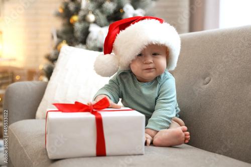 Cute baby in Santa hat with Christmas gift sitting on sofa at home © New Africa