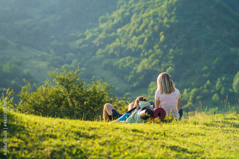 Young Caucasian couple hiking in a wild mountain area sitting on the grass and looking away into the green forests on a sunny summer day.