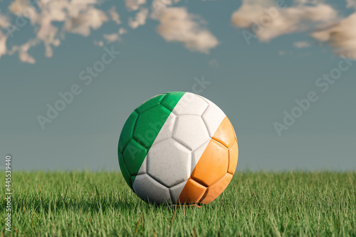 Soccer ball with the national colors of Ireland on a green meadow. Leather in slightly used look. Background blue with clouds. 3D illustration.