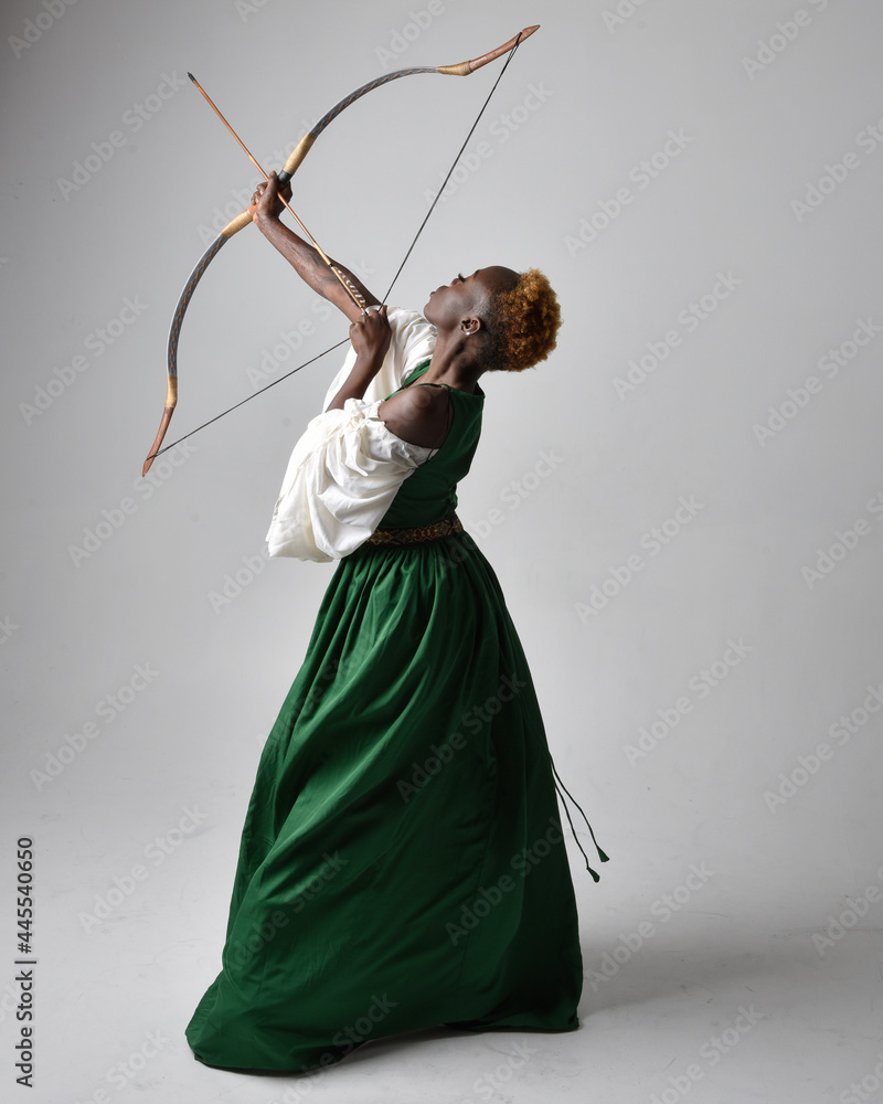 Full length portrait of girl with red hair wearing brown medieval archer  costume.. Standing pose with back to the camera holding a bow and arrow,  isolated against a grey studio background. Stock