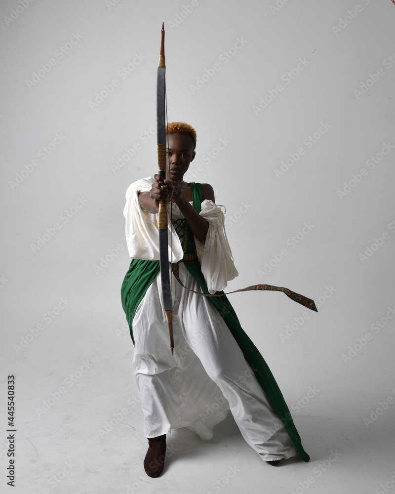 Full length portrait of pretty African woman wearing long green medieval fantasy gown holding bow and arrow, standing action pose on a light grey studio background.