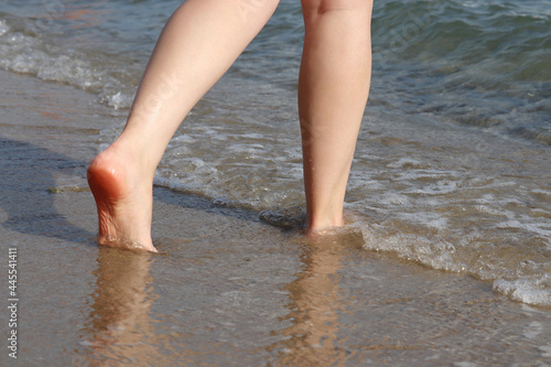 Barefoot woman walking by the sand to the sea waves. Naked female legs in water, beach vacation