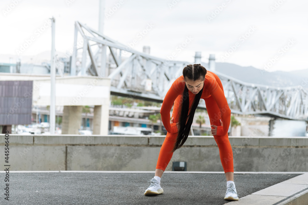 Young athlete stop to rest while running. Copy space. Tired woman bending over to catch her breath jogging in modern city.