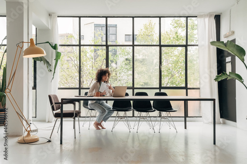 Young mixed race female student showing notes to a friend in a video call. Spacious loft apartment living room. copy space.