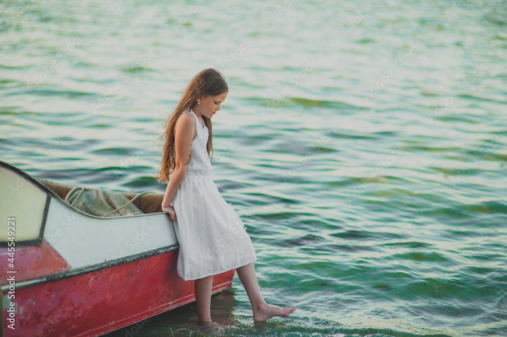 Pin-up girl teenager with long hair in a white dress stands leaning against a fishing boat in the sea water