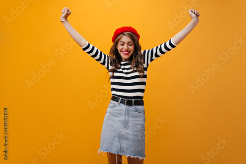 Cheerful woman in beret and shirt happily posing on orange background. Positive cute girl with curly hair and red lips posing.. © Look!