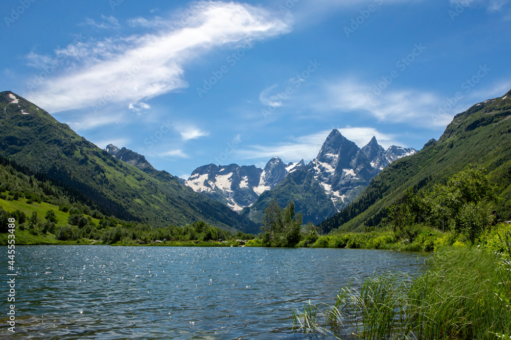 Landscape with a mountain lake on a summer day. Blue sky with clouds. Grass in the foreground. Mountain snow peaks.