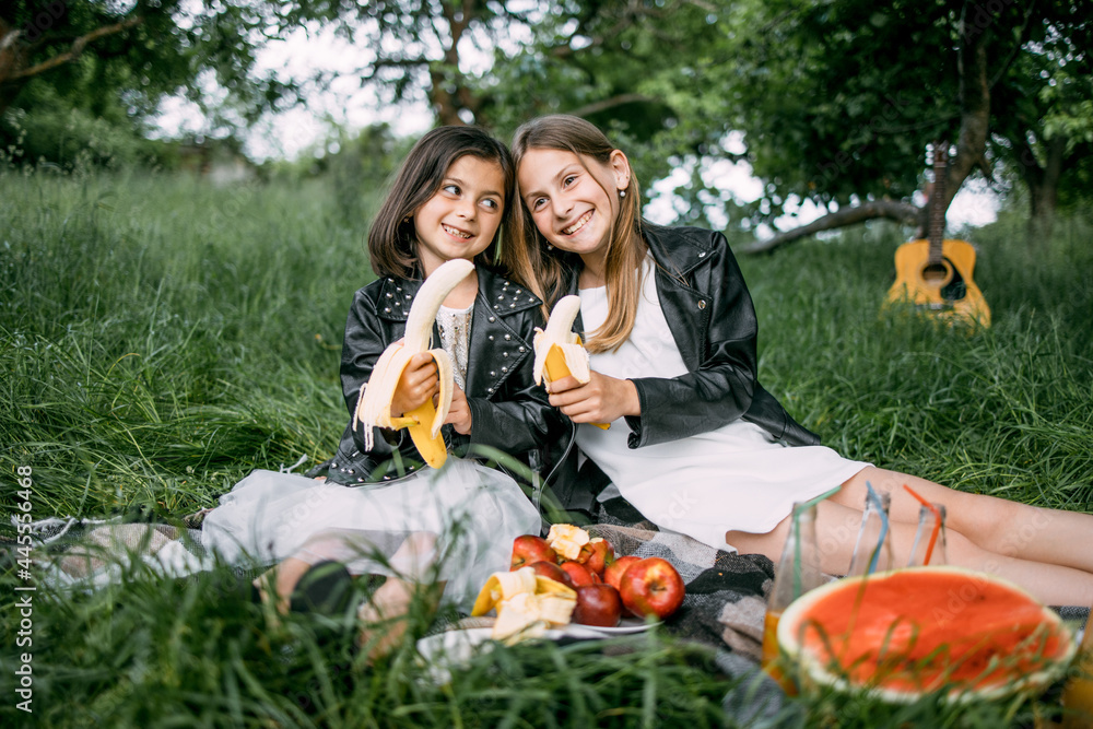 Fototapeta premium Cute happy little sisters sitting closely to each other, holding bananas in hands and smiling sincerely on camera. Charming kids spending time together on fresh air.