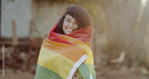Young curly hair woman covering with lgbt pride flag. Alone. One. Keeping fist up, covering LGBT flag. LGBT+ flag on outdoor background.