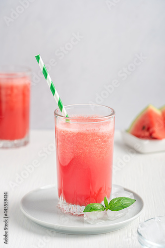 Red refreshing cold watermelon smoothie drink served in drinking glass with ice cubes and paper straw on round plate with ripe fruit slices and basil leaf on white wooden table ib hot summer season