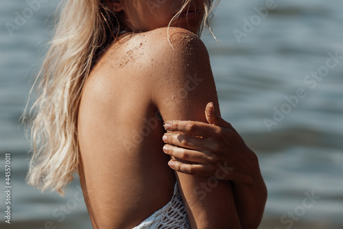 Obraz na plátně Cropped shot of a young tanned slim blonde woman in a white knitted top touches her arm and scrubs the skin with sand on the background of the sea