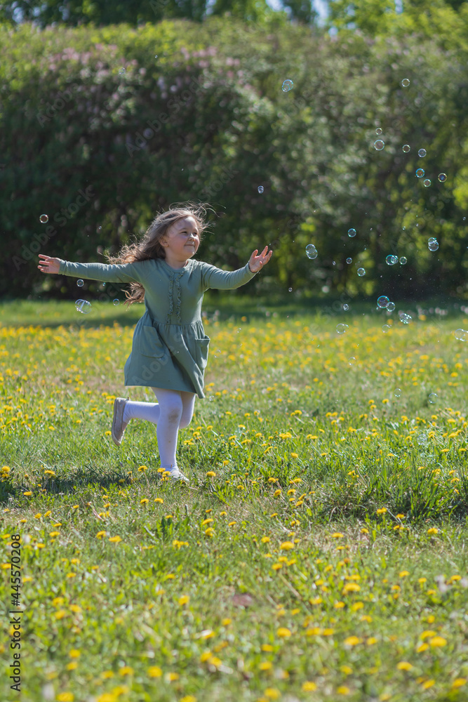 A little girl with long curly blond hair in a green dress on a yellow meadow catches soap bubbles. Image with selective focus. A little girl with long curly blond hair in a green dress on a yellow meadow catches soap bubbles. Image with selective focus.