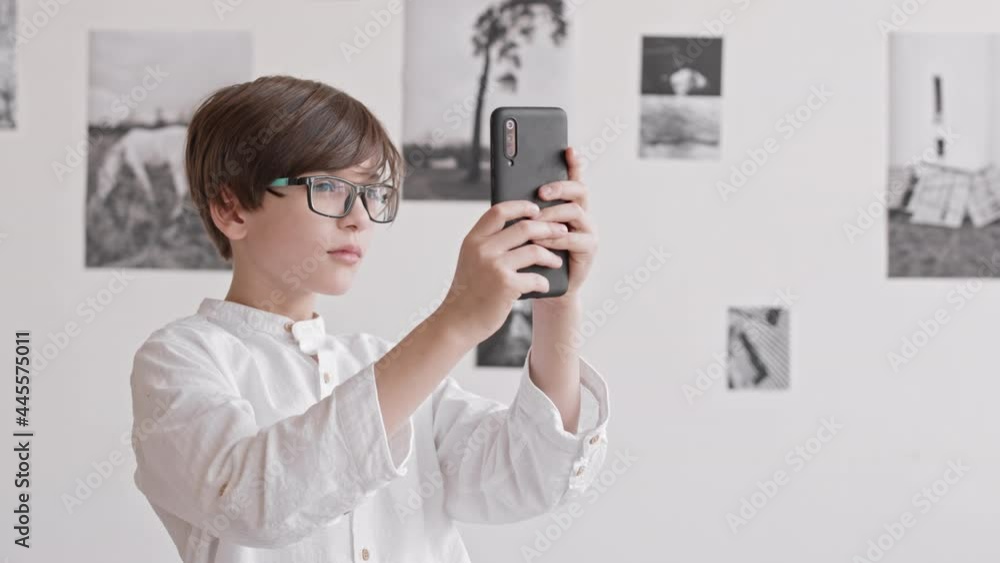 Waist-up of Caucasian boy wearing glasses standing in museum at contemporary photography exhibition, talking pictures with smartphone, turning around Waist-up of Caucasian boy wearing glasses standing in museum at contemporary photography exhibition, talking pictures with smartphone, turning around