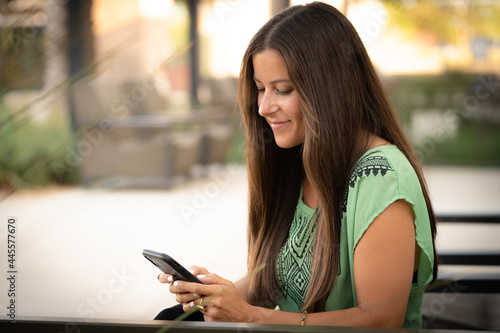 Beautiful Pretty Young Brunette Woman Texting On Smartphone Phone Cell Phone Outside on at Mall Café with Natural Soft Daylight.