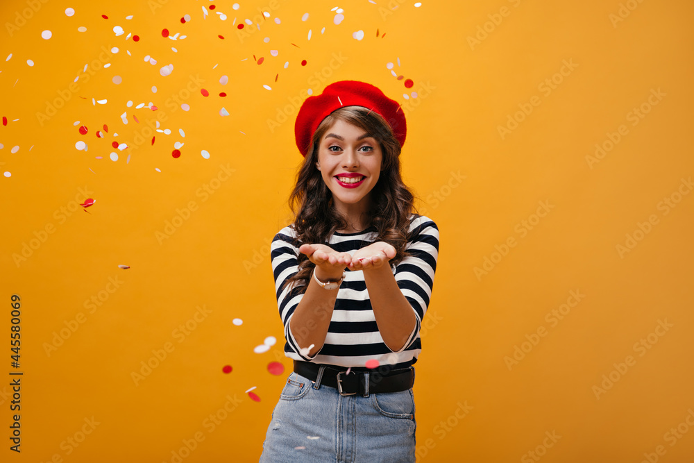 Attractive lady in red beret and shirt throwing confetti. Charming young woman with bright lips posing on isolated background.. Attractive lady in red beret and shirt throwing confetti. Charming young woman with bright lips posing on isolated background..