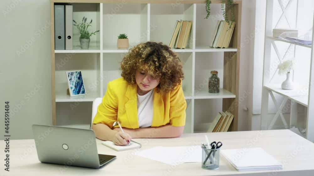 Remote education. Online courses. Distance learning. Home office. Young concentrated curvy student girl with curly hair taking notes in light interior room. Remote education. Online courses. Distance learning. Home office. Young concentrated curvy student girl with curly hair taking notes in light interior room.